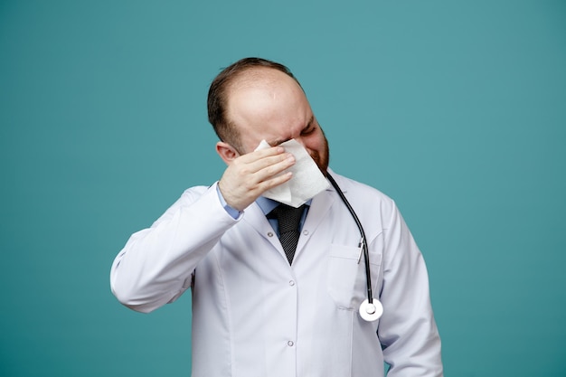 Unhealthy young male doctor wearing medical coat and stethoscope around his neck wiping his eye with napkin with closed eyes isolated on blue background