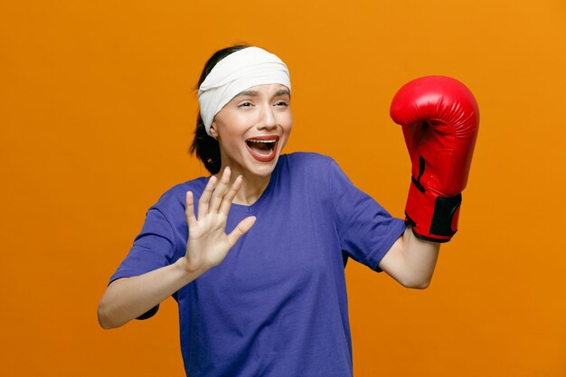 Unhealthy young sporty woman wearing tshirt and boxing glove looking at side showing hi gesture with head wrapped with bandage isolated on orange background