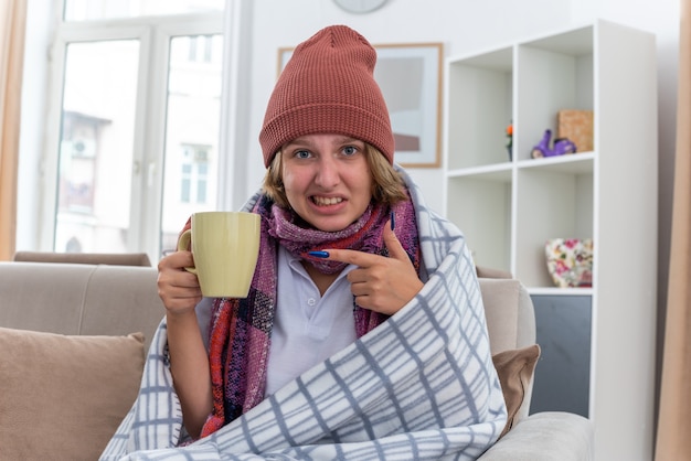 Giovane donna malsana in cappello con sciarpa calda intorno al collo che beve tè caldo per ottenere una migliore sofferenza da raffreddore e influenza seduto sul divano nel soggiorno luminoso