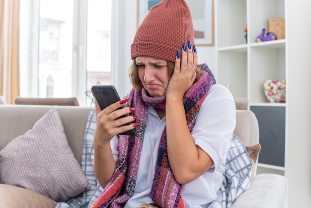 Free photo unhealthy young woman in hat with warm scarf around neck feeling unwell and sick suffering from cold and flu having strong headache and fever holding smartphone sitting on couch in light living room