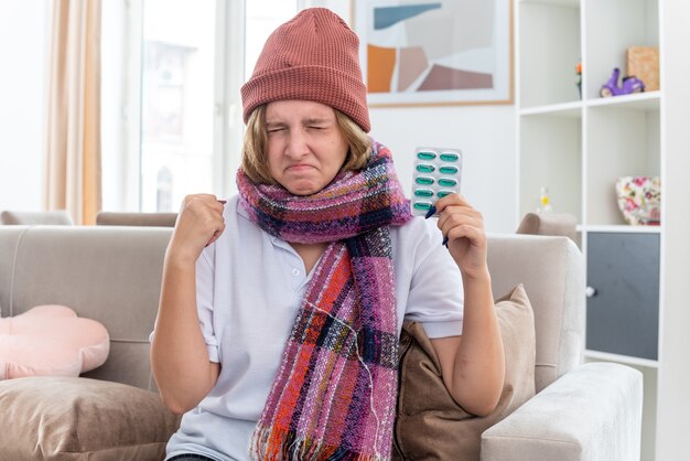 Unhealthy young woman in hat with warm scarf around neck feeling unwell and sick suffering from cold and flu holding pills clenching fist eyes closed sitting on couch in light living room