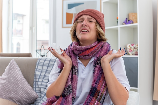 Unhealthy young woman in hat with warm scarf around neck feeling unwell and sick suffering from cold and flu looking worried and disappointed with arms raised sitting on couch in light living room