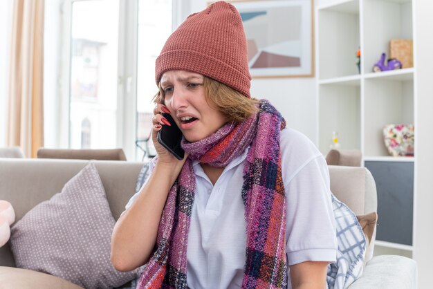 Unhealthy young woman in hat with warm scarf around neck feeling unwell and sick suffering from cold and flu looking worried and sad while talking on mobile phone sitting on couch in light living room