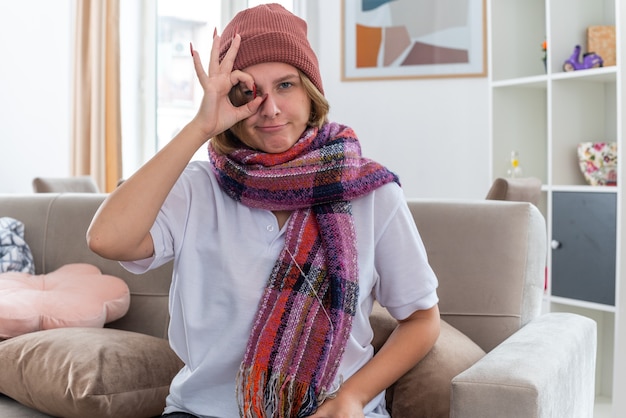 Foto gratuita giovane donna malsana in cappello con sciarpa calda intorno al collo che soffre di raffreddore e influenza sentirsi meglio facendo segno ok sorridente seduta sul divano in un soggiorno luminoso