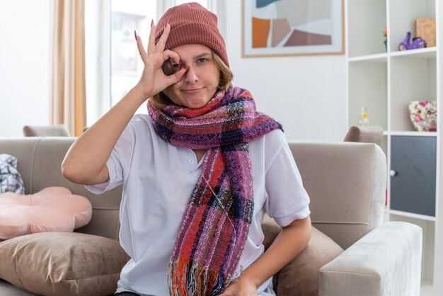 Unhealthy young woman in hat with warm scarf around neck suffering from cold and flu feeling better doing ok sign smiling sitting on couch in light living room