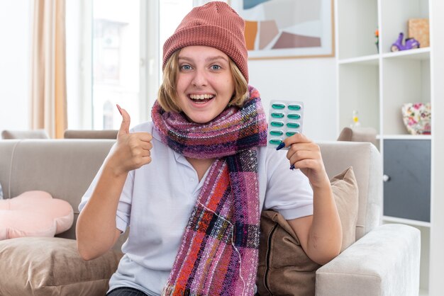 Unhealthy young woman in hat with warm scarf around neck suffering from cold and flu holding pills showing thumbs up smiling feeling better sitting on couch in light living room