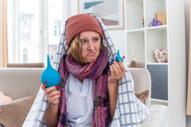 Unhealthy young woman in hat wrapped in blanket feeling unwell and sick holding enemas looking confused having doubts sitting on couch in light living room