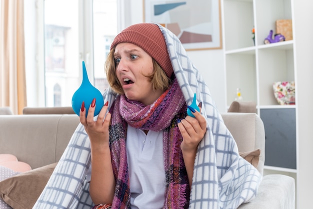 Unhealthy young woman in hat wrapped in blanket feeling unwell and sick holding enemas looking confused sitting on couch in light living room