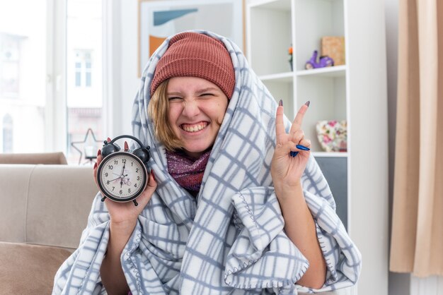 Unhealthy young woman in hat wrapped in blanket holding alarm clock  smiling showing v-sign feeling better sitting on couch in light living room