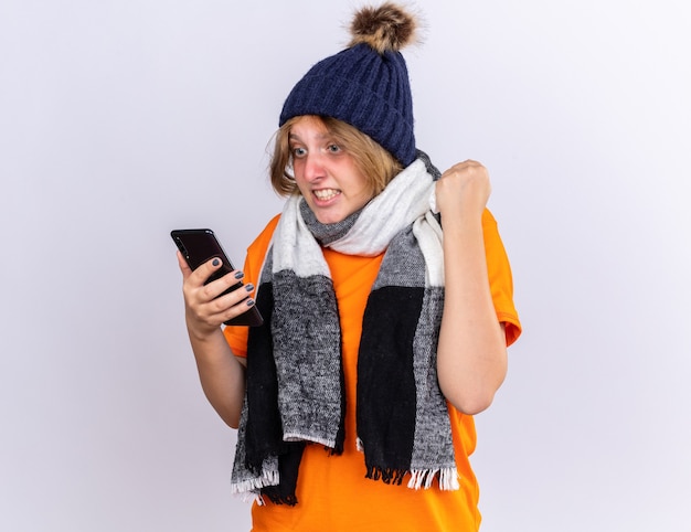 Нездоровая молодая женщина в оранжевой футболке с теплым шарфом на шее и шляпе чувствует себя ужасно, глядя на свой смартфон с агрессивным выражением лица, поднимая кулак