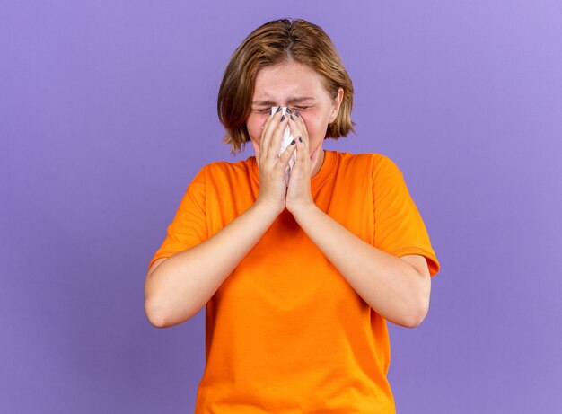 Unhealthy young woman in orange t-shirt feeling terrible blowing running nose caught cold sneezing in tissue standing over purple wall