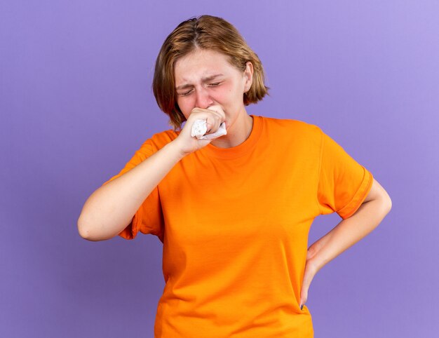 Нездоровая молодая женщина в оранжевой футболке, чувствуя ужасный кашель в кулаке, страдает от вируса, стоящего над фиолетовой стеной