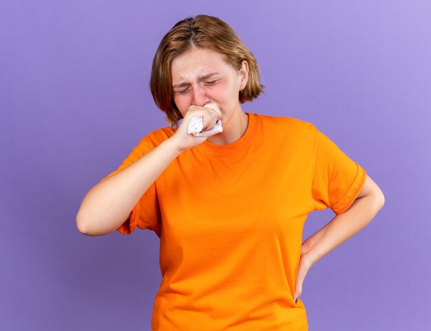 Unhealthy young woman in orange t-shirt feeling terrible coughing in fist suffering from virus standing over purple wall