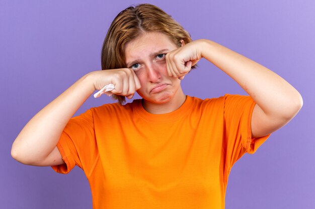 Unhealthy young woman in orange t-shirt feeling terrible holding tissue suffering from running nose cought cold crying hard rubbing eyes standing over purple wall