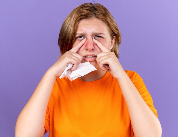 Unhealthy young woman in orange t-shirt feeling terrible holding tissue suffering from running nose cought cold