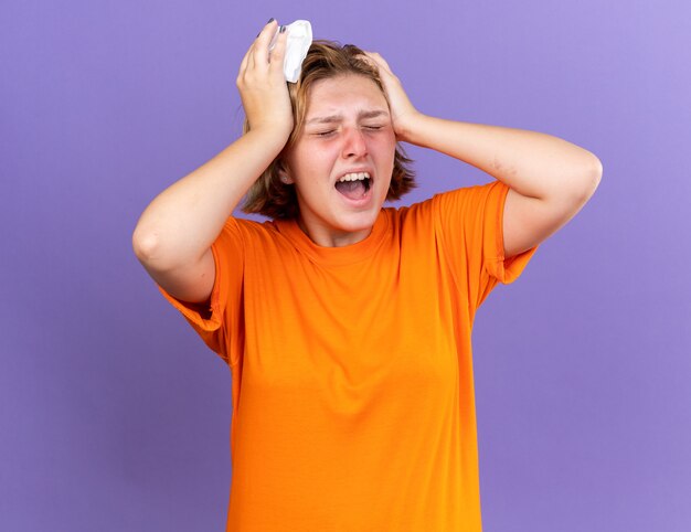 Нездоровая молодая женщина в оранжевой футболке чувствует себя ужасно крича, касаясь ее головы, страдающей от лихорадки и сильной головной боли