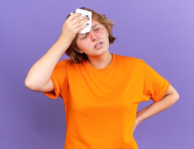 Foto gratuita giovane donna malsana in maglietta arancione che si sente terribile toccarsi la testa mentre si sente stordito avendo l'influenza che soffre di forte mal di testa in piedi sul muro viola