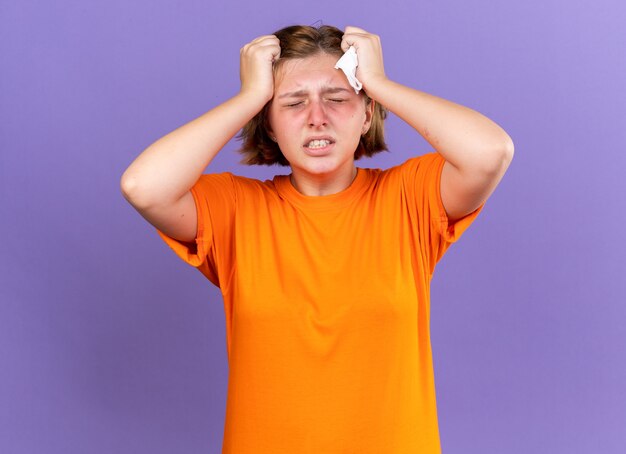 Нездоровая молодая женщина в оранжевой футболке чувствует себя ужасно, касаясь ее головы, когда у нее кружится голова от гриппа, страдающего сильной головной болью