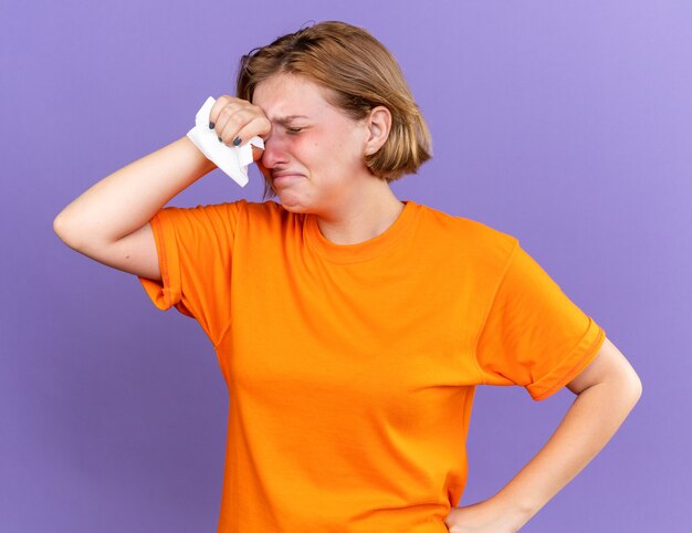 Unhealthy young woman in orange t-shirt feeling terrible with tissue touching her forehead having fever and headache suffering from virus