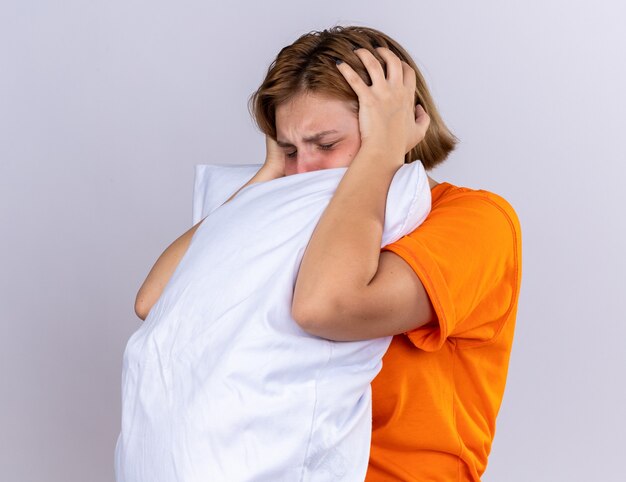 Unhealthy young woman in orange t-shirt holding pillow feeling sick suffering from flu having fever and strong headache standing over white wall