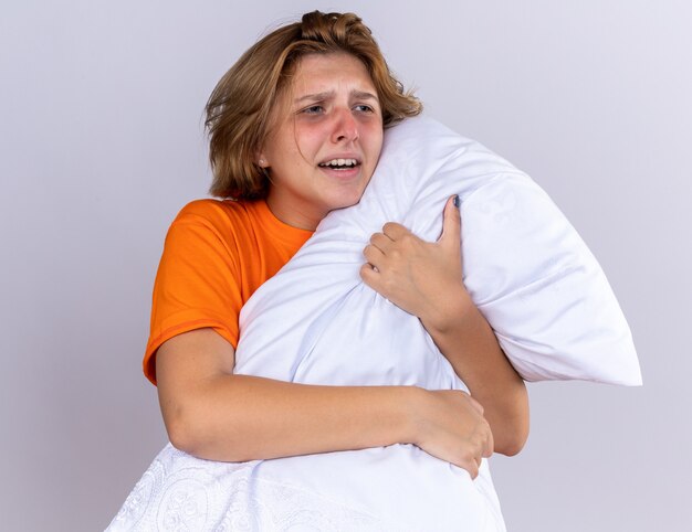 Нездоровая молодая женщина в оранжевой футболке, держащая подушку, чувствует себя больной гриппом, стоя над белой стеной