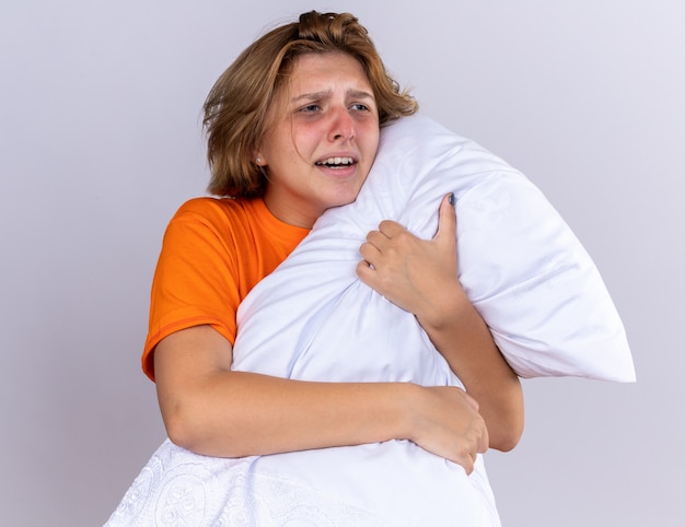 흰 벽 위에 서있는 독감으로 고통받는 아픈 느낌의 베개를 들고 오렌지 티셔츠에 건강에 해로운 젊은 여자