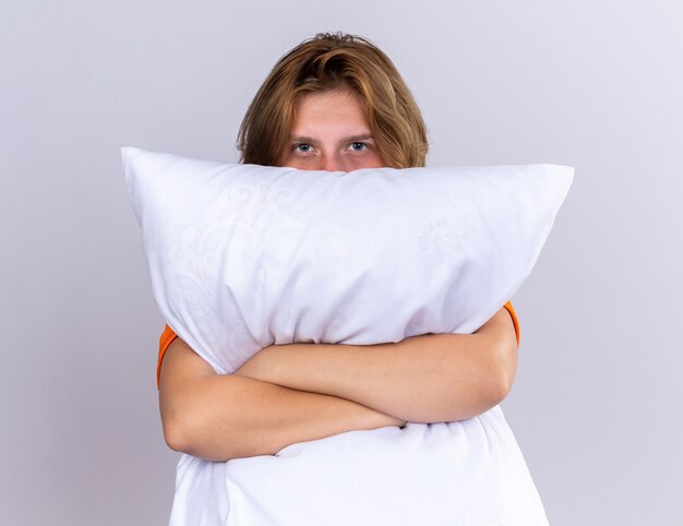 Unhealthy young woman in orange t-shirt holding pillow feeling unwell covering her face peeking over pillow standing over white wall