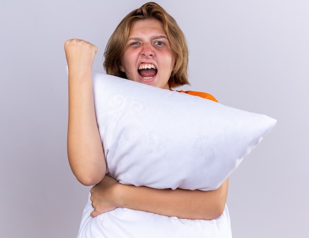 Unhealthy young woman in orange t-shirt holding pillow feeling unwell peeking over pillow shouting with aggressive expression showing her fist standing over white wall