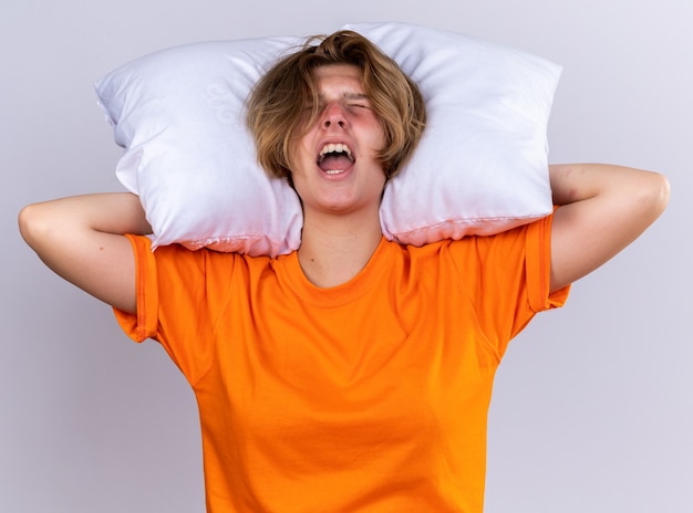 Giovane donna malsana in maglietta arancione che tiene il cuscino che si sente male gridando con un'espressione aggressiva in piedi sul muro bianco white