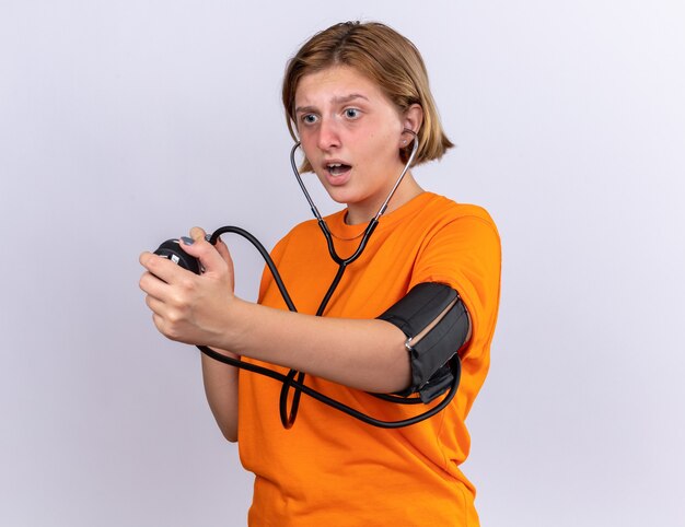 Unhealthy young woman in orange t-shirt measuring blood pressure using tonometer looking worried