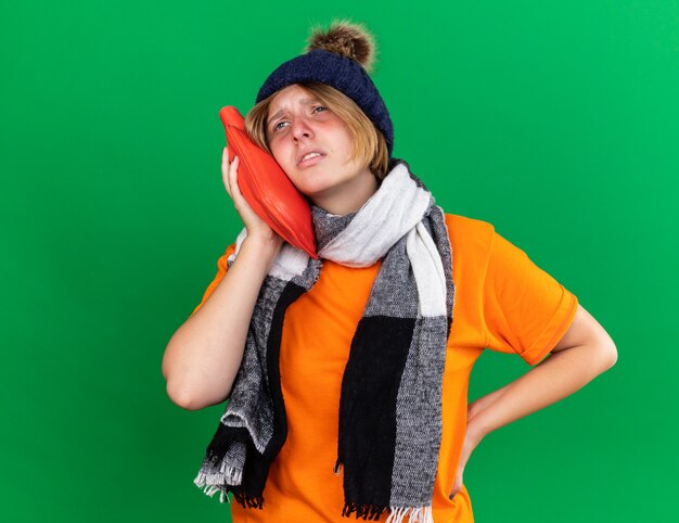 Unhealthy young woman in orange t-shirt with hat and warm scarf around neck feeling terrible holding hot-water bottle suffering from cold