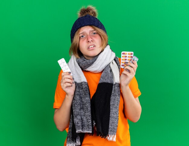 독감으로 고통받는 끔찍한 약을 들고 목에 따뜻한 스카프와 모자가 달린 주황색 티셔츠에 건강에 해로운 젊은 여성