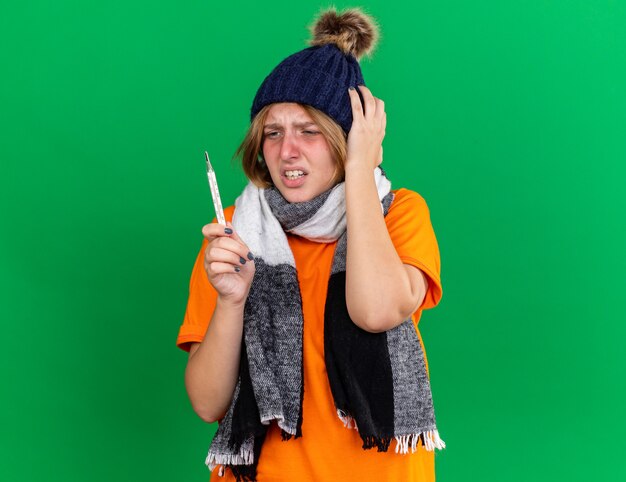 독감으로 고통받는 열이있는 끔찍한 지주 온도계 느낌 모자와 따뜻한 스카프가 달린 주황색 티셔츠에 건강에 해로운 젊은 여성