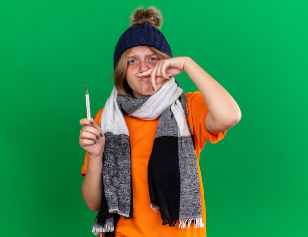 Unhealthy young woman in orange t-shirt with hat and warm scarf around neck feeling terrible holding thermometer having fever suffering from running nose standing over green wall