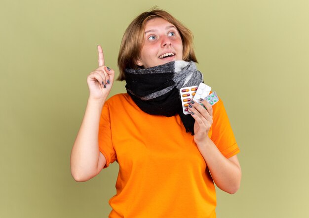 Unhealthy young woman in orange t-shirt with warm scarf around neck feeling better holding different pills smiling having new idea showing index finger