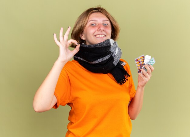 Нездоровая молодая женщина в оранжевой футболке с теплым шарфом на шее чувствует себя лучше, держа разные таблетки, улыбаясь, показывая знак ОК