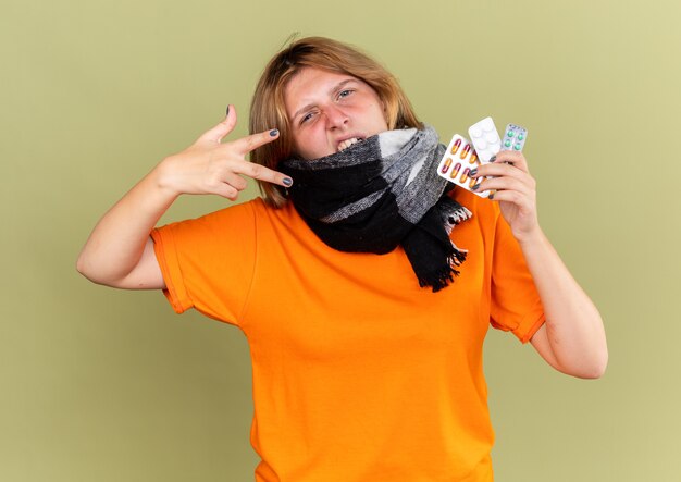 Unhealthy young woman in orange t-shirt with warm scarf around neck feeling better suffering from flu holding different pills looking confident