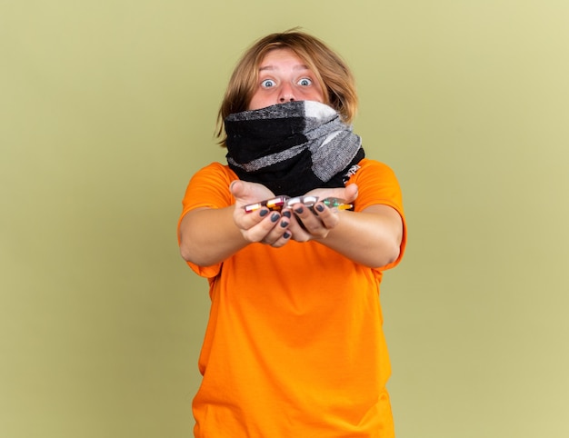 Free photo unhealthy young woman in orange t-shirt with warm scarf around neck feeling terrible suffering from flu holding different pills looking worried standing over green wall