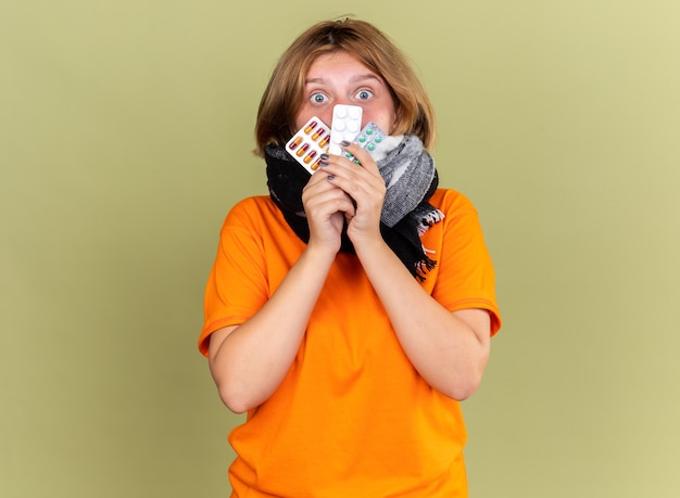 Free photo unhealthy young woman in orange t-shirt with warm scarf around neck feeling unwell suffering from flu holding different pills looking confused and worried standing over green wall