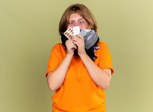Нездоровая молодая женщина в оранжевой футболке с теплым шарфом на шее чувствует недомогание, страдает от гриппа, держит в руках разные таблетки и выглядит смущенной