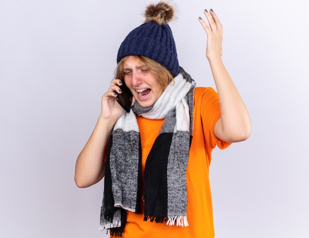 목과 모자 주위에 따뜻한 스카프가 달린 주황색 티셔츠에 건강에 해로운 젊은 여성이 흰 벽에 걱정되는 공격적인 표정으로 휴대 전화로 이야기하는 독감으로 끔찍한 고통을 느낍니다.