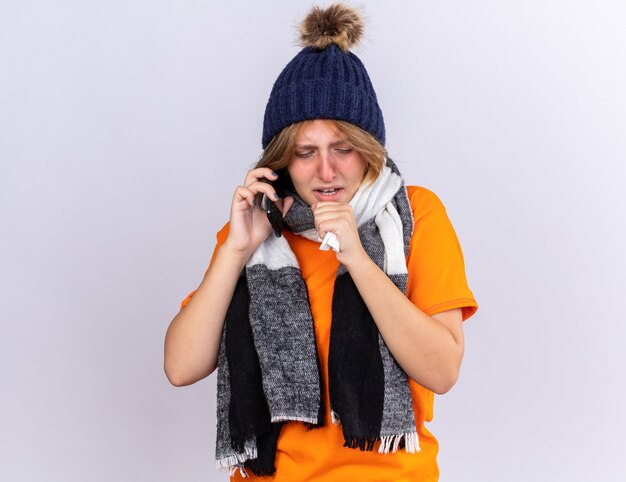 Unhealthy young woman in orange t-shirt with warm scarf around neck and hat feeling terrible talking on mobile phone blowing running nose sneezing in tissue