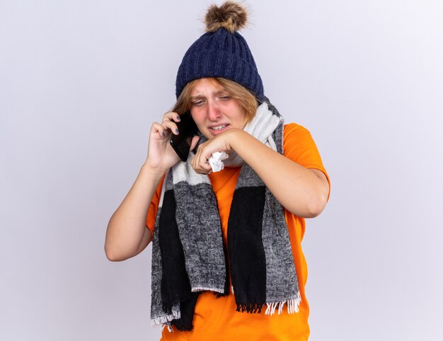 オレンジ色のTシャツを着た不健康な若い女性。首に暖かいスカーフを巻いて、帽子をかぶって携帯電話で話すのがひどい。鼻水が組織にくしゃみをしている。