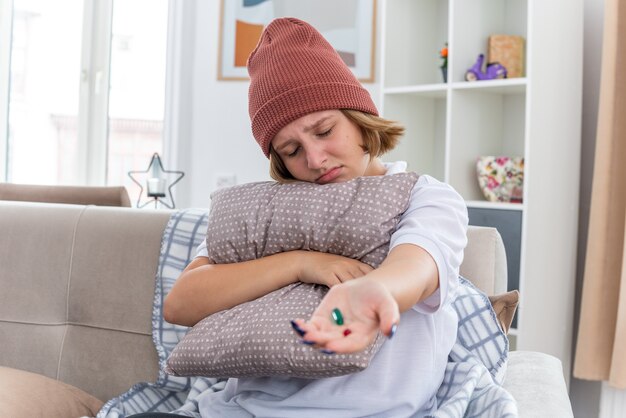 Unhealthy young woman in warm hat with blanket feeling unwell suffering from cold and flu holding pillow and pills upset and sick sitting on couch in light living room