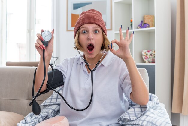 Unhealthy young woman in warm hat with blanket  surprised doing ok sign checking her blood pressure using tonometer sitting on couch in light living room