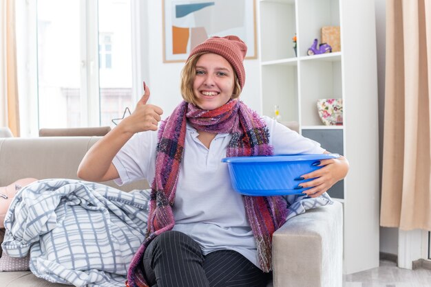 Unhealthy young woman in warm hat with scarf around neck holding basin feeling nauseous looking smiling showing thumbs up feeling better sitting on couch in light living room