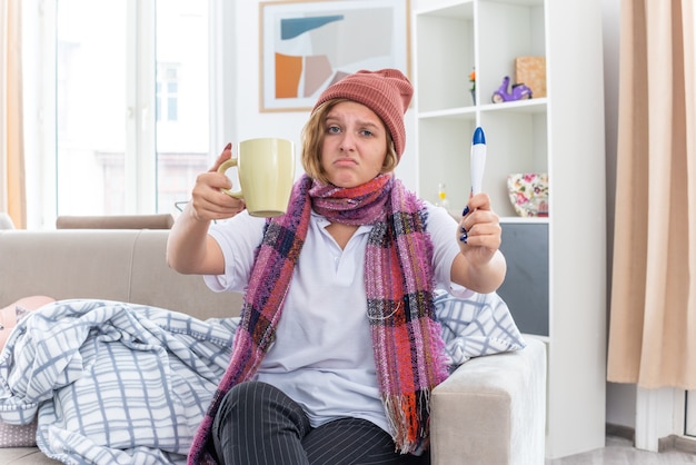Нездоровая молодая женщина в теплой шляпе с шарфом на шее, держащая чашку чая и термометр, выглядит обеспокоенной, сидя на диване в светлой гостиной