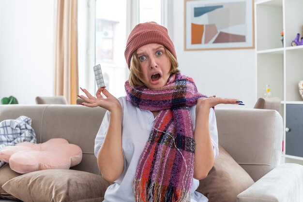 Unhealthy young woman in warm hat with scarf around neck holding feeling unwell and sick suffering from cold and flu holding pills spreading arms to the sides sitting on couch in light living room