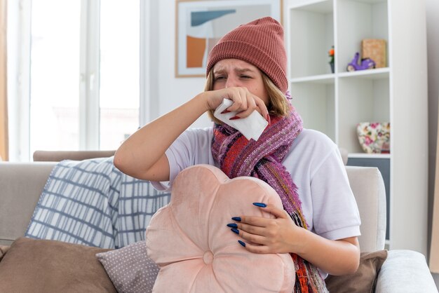 Unhealthy young woman in warm hat with scarf blowing nose in tissue sneezing suffering from cold and flu sitting on the chair in light living room