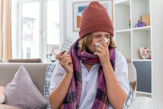 Unhealthy young woman in warm hat with scarf blowing nose in tissue sneezing suffering from cold and flu sitting on the chair in light living room